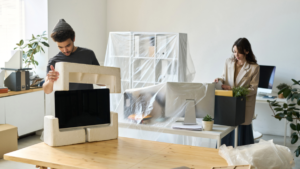 Male packing computer, female packing in a box. Wrapped office furniture in plastic