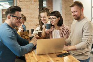 a group of office workers meeting around a table, drinking coffee and looking at a laptop